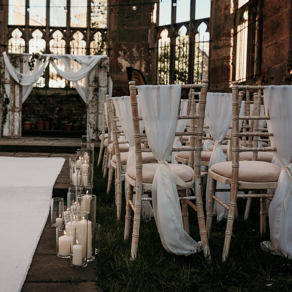 Outdoor wedding ceremony at St Lukes Bombed Out Church in Liverpool with chiffon chair back covers and pillar candles lining the aisle 
