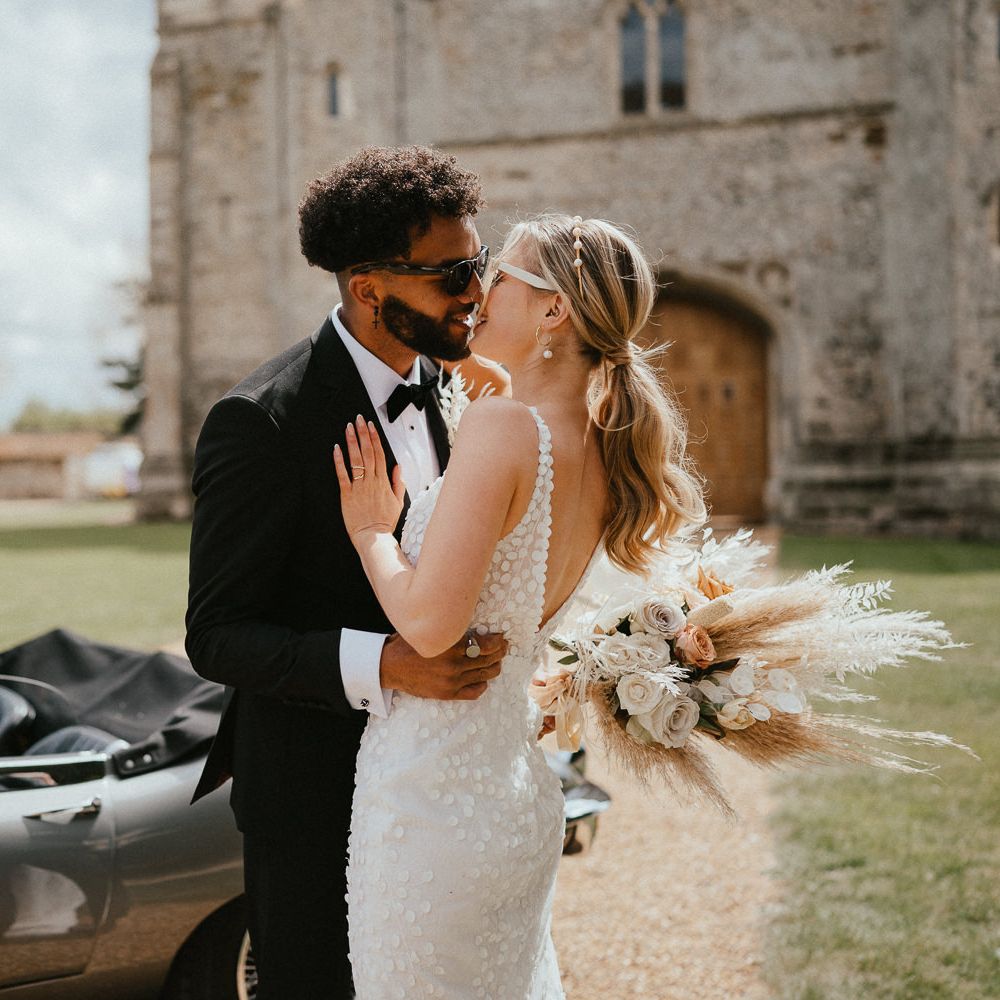 Groom in a tuxedo kissing his bride in an appliue wedding dress with sleek ponytail and dried flower bouquet outside Pentney Abbey, Norfolk