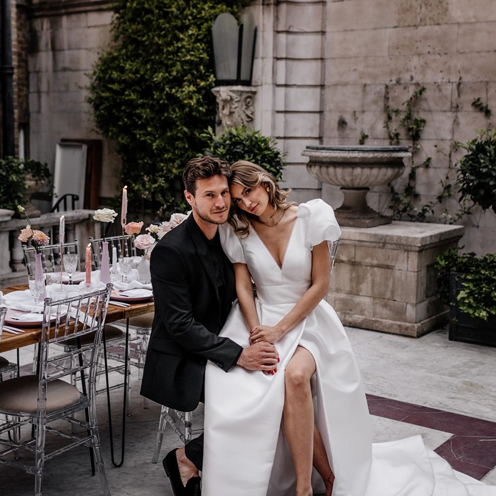 Bride in a puff sleeve wedding dress with front split sitting on her grooms lap at their stylish outdoor wedding reception 