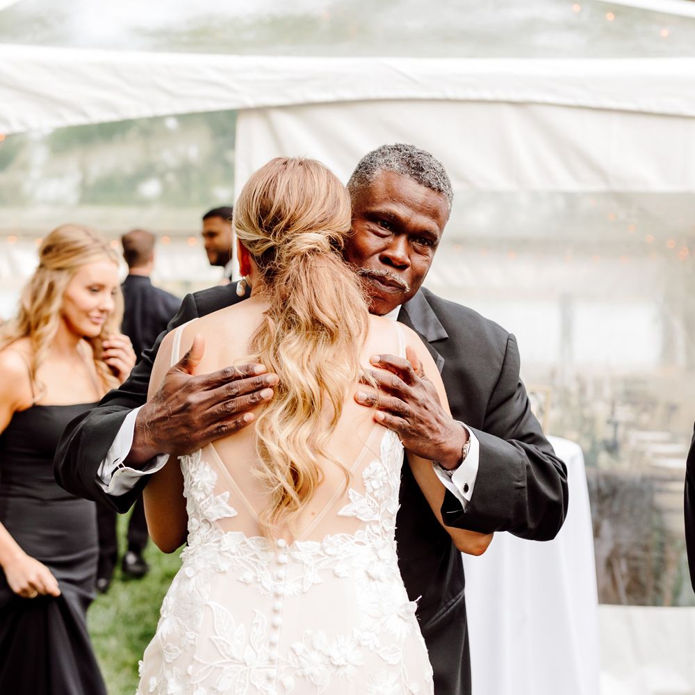 Bride in embroidered low back Made With Love wedding dress and curled ponytail hugs man in black suit outside marquee at backyard wedding
