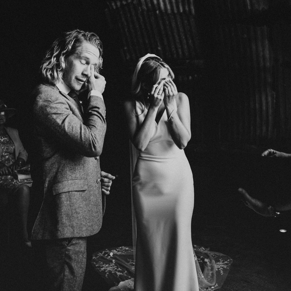 Bride in white Made With Love wedding dress and veil and groom in three piece suit both wipe eyes during wedding ceremony in hut at Anran Devon