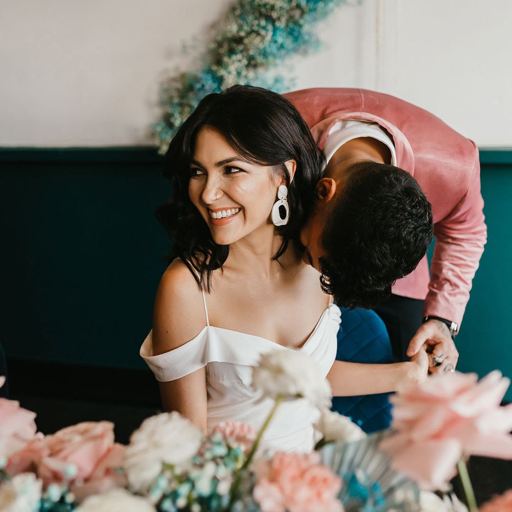 Groom in a pink velvet jacket kissing his bride on the shoulder with short curly hair wearing a cold shoulder wedding dress and dangly earrings 