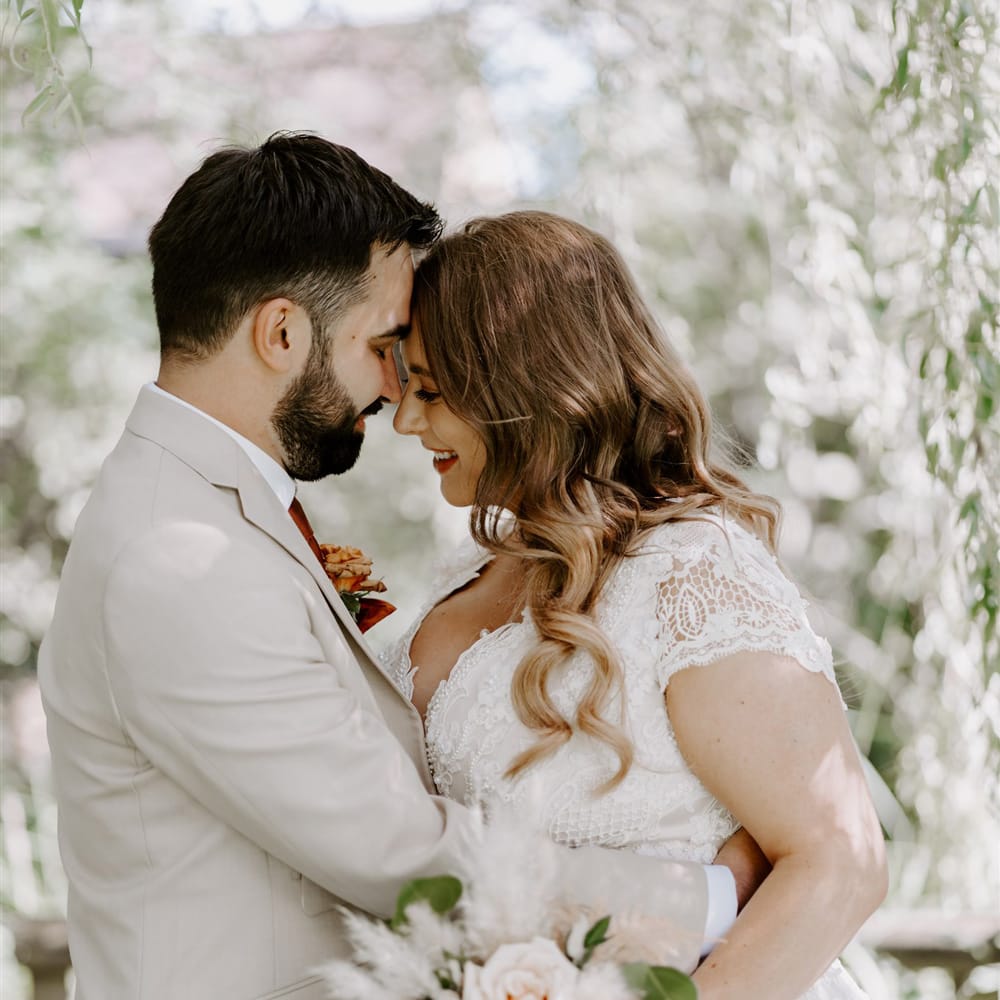 Bride in homemade lace wedding dress with mixed rose bridal bouquet and curled hair hugs groom in beige suit with floral buttonhole at DIY garden wedding ceremony