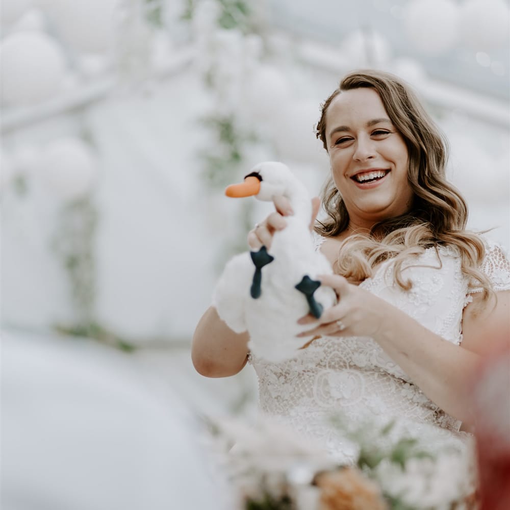 Bride in lace homemade wedding dress and curled hair holding toy swan smiles during marquee wedding reception 
