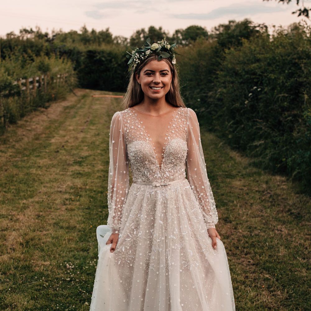 Bride in long mesh sleeve embroidered Jule Vino wedding dress with v neck and boho flower crown walks through field after outdoor wedding reception
