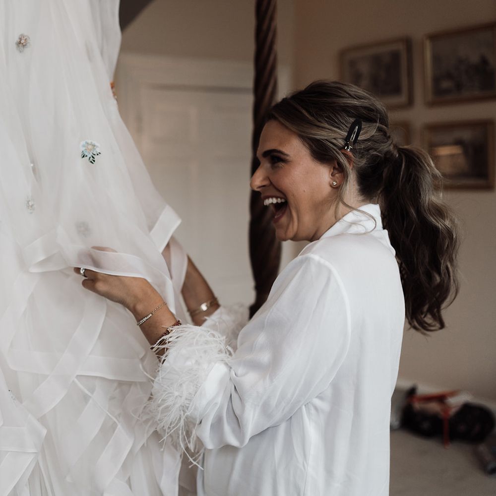 Bride in white fur trim satin robe stands holding train of embroidered Victoria Sanders wedding dress