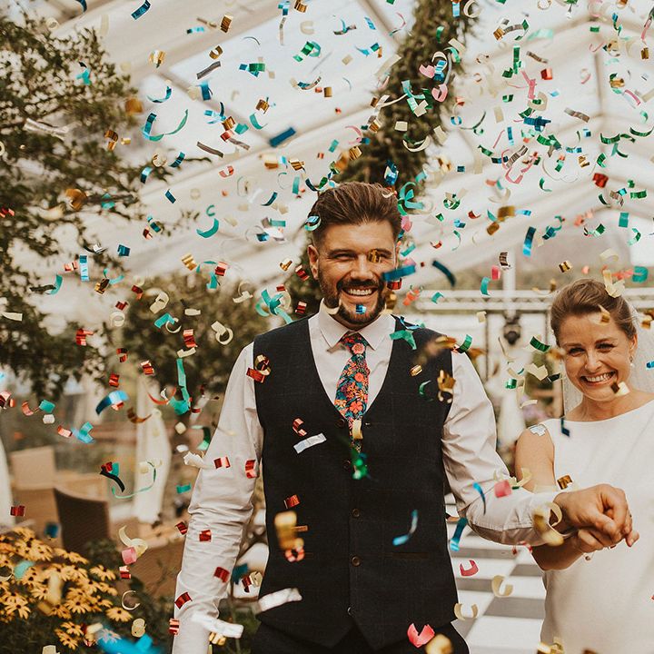 Colourful confetti moment in a clear marquee with groom in a navy waistcoat and floral tie and bride in a slip wedding dress 