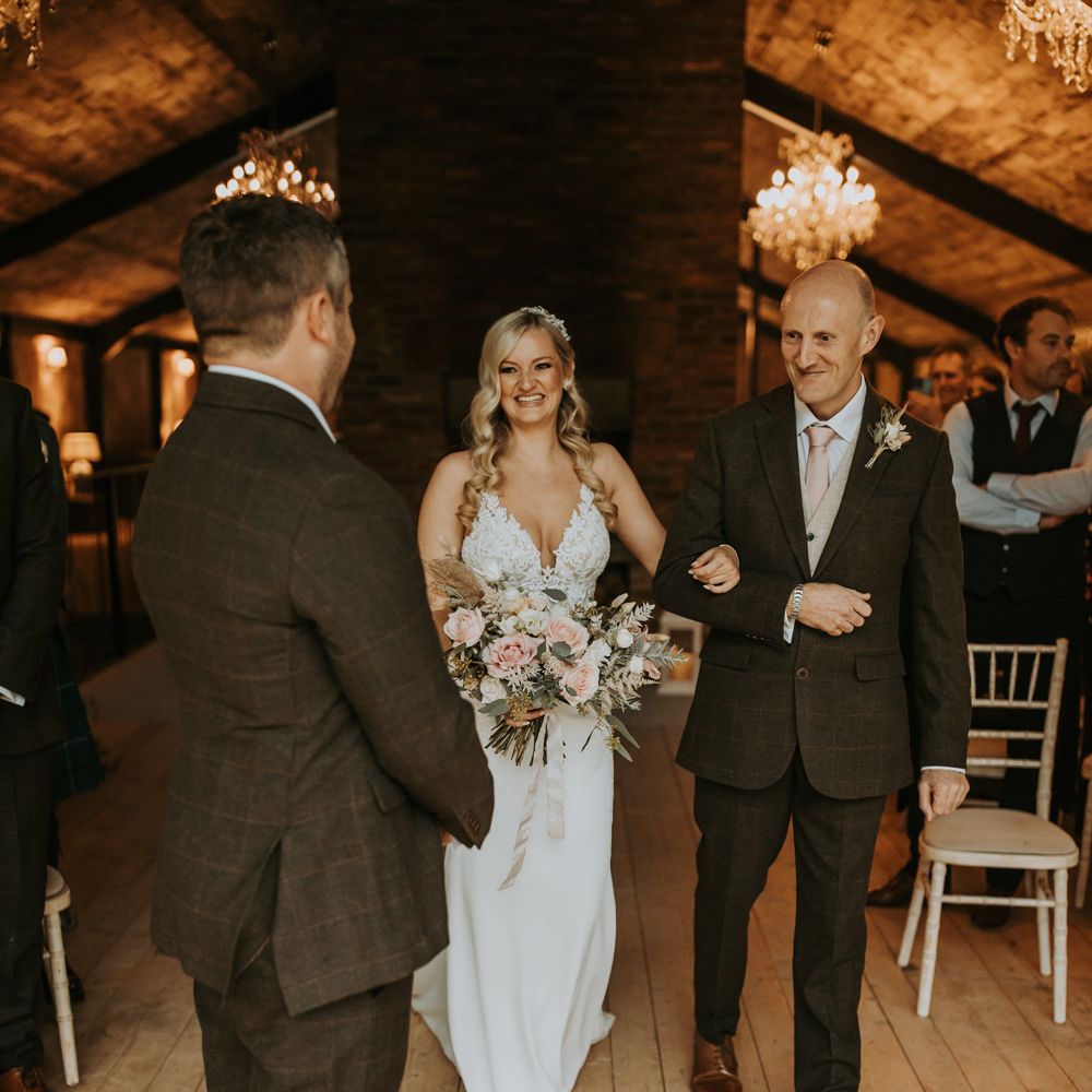 Bride with curled blonde hair and lace wedding dress holding white, green and pink bridal bouquet smiles as she walks arm in arm with father of the bride down barn aisle