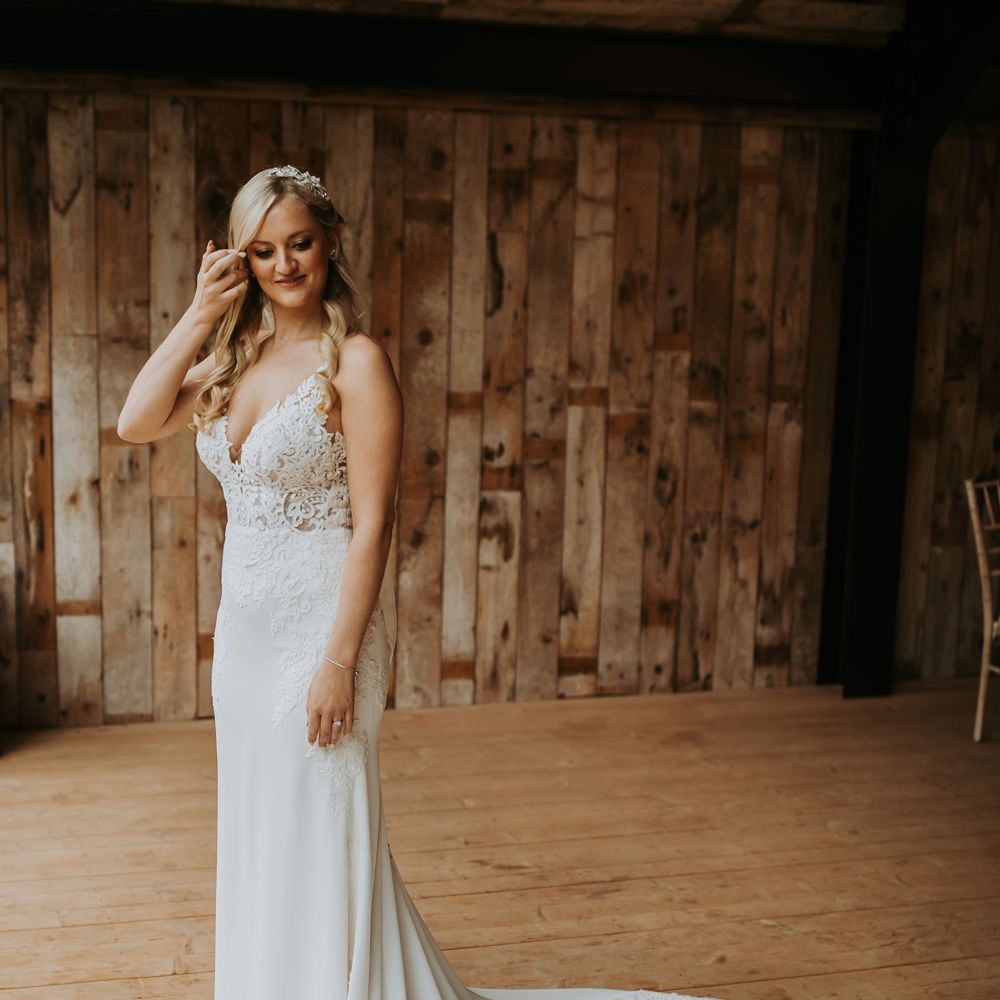 Bride in lace wedding dress with train stands by window at Hidden River Barn wedding venue in the Lake District