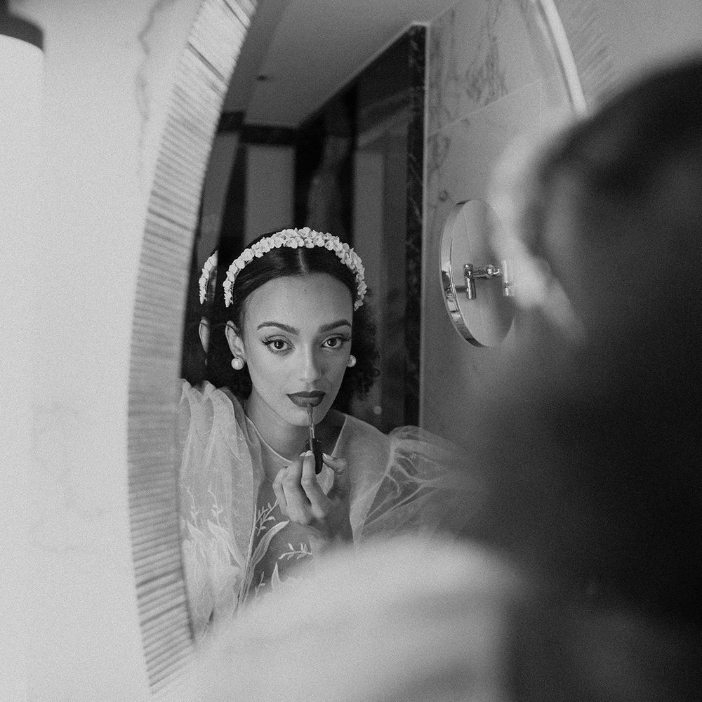 Black bride with sleek back hair wearing an appliqué headband looking in the mirror applying her lipstick 