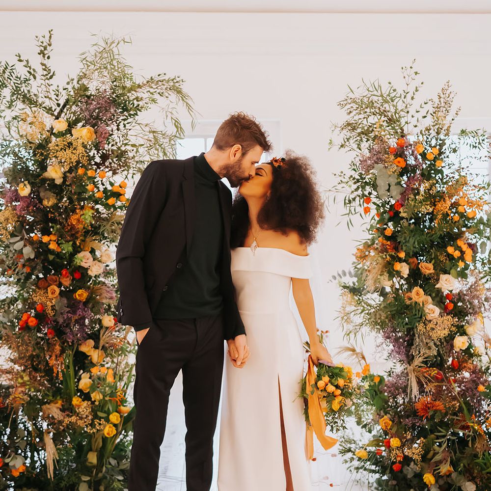 Bride in an off the shoulder wedding dress with side slit and groom in all black kissing between column golden wedding ceremony flowers