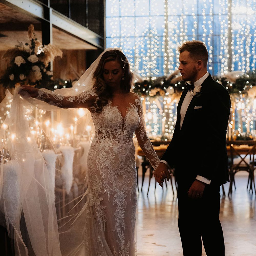 Bride in lace wedding dress holds out her veil as groom looks at her mesmerised in front of fairy lights