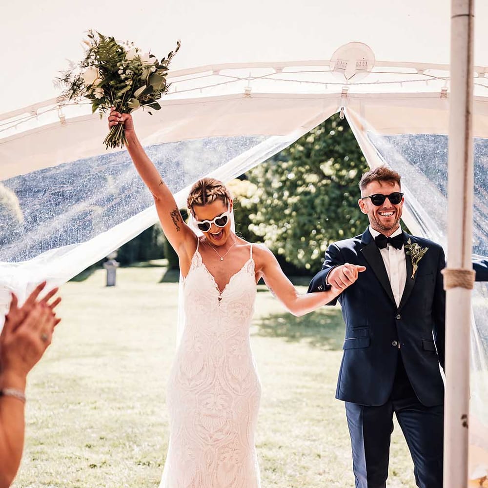 Groom in blue tuxedo with black sunglasses walks in with the bride in lace wedding dress with retro heart sunglasses 