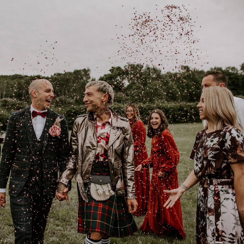 Grooms walk through confetti outdoors as wedding guests surround them as groom wears kilt and gold leather jacket