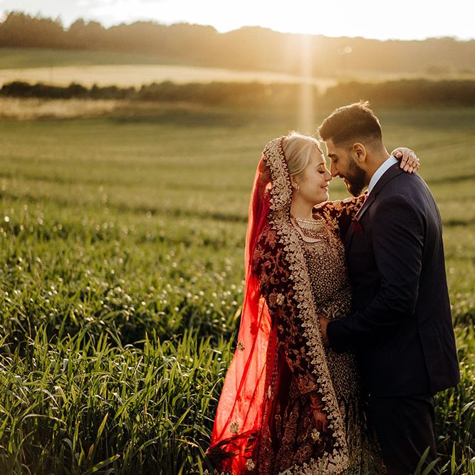 Bride wearing brightly coloured saree and her groom in deep blue three piece suit stand in field during golden hour for couples portraits