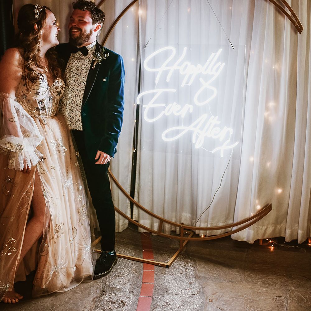 Bride & groom stand beside neon wedding sign that hangs within moon shaped stand in front of curtain 