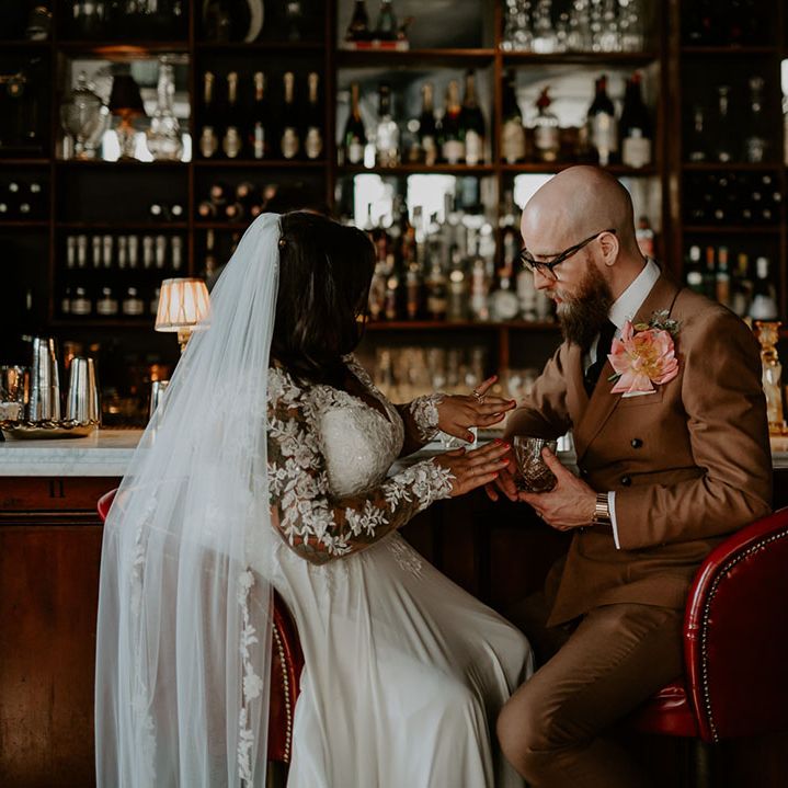 Bride & groom in retro styled brown suit with statement pink floral buttonhole sit at bar drinking whiskey 