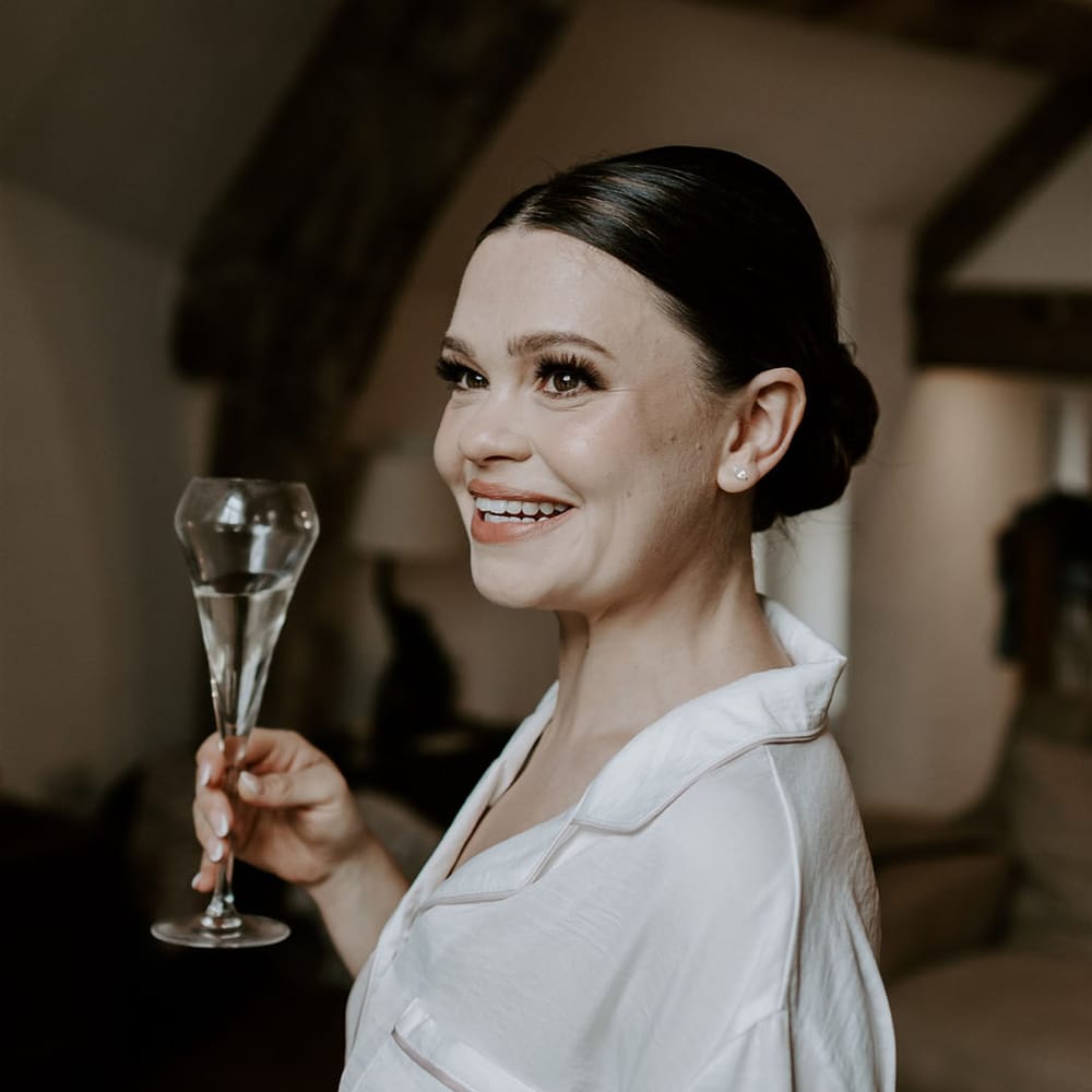 Bride drinks champagne as she gets ready for the wedding in white pyjamas with pink trim and hair in a sleek bun 