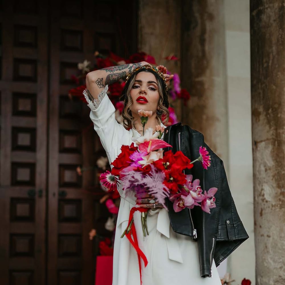 Bride holding bridal bouquet with hot pink, bright pink and viva magenta dahlias, orchids, roses, amaryllis, anthuriums and dried flowers wearing blazer style wrap around short wedding dress with delicate fringe detailing with black leather jacket on her shoulder