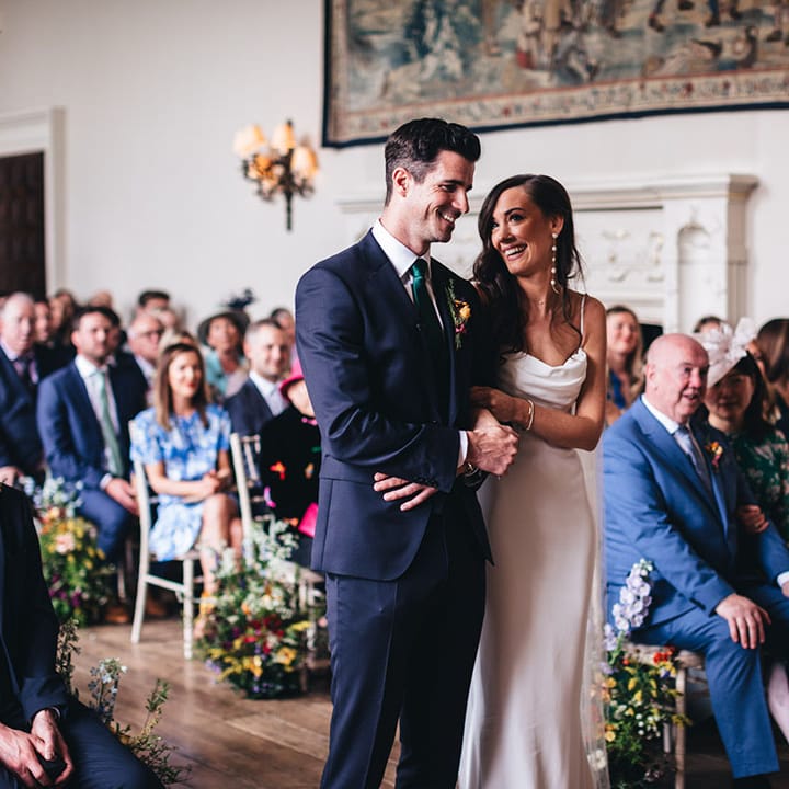 Groom in a green and blue suit stands linking arms with the bride smiling as they participate in their civil wedding ceremony at Elmore Court