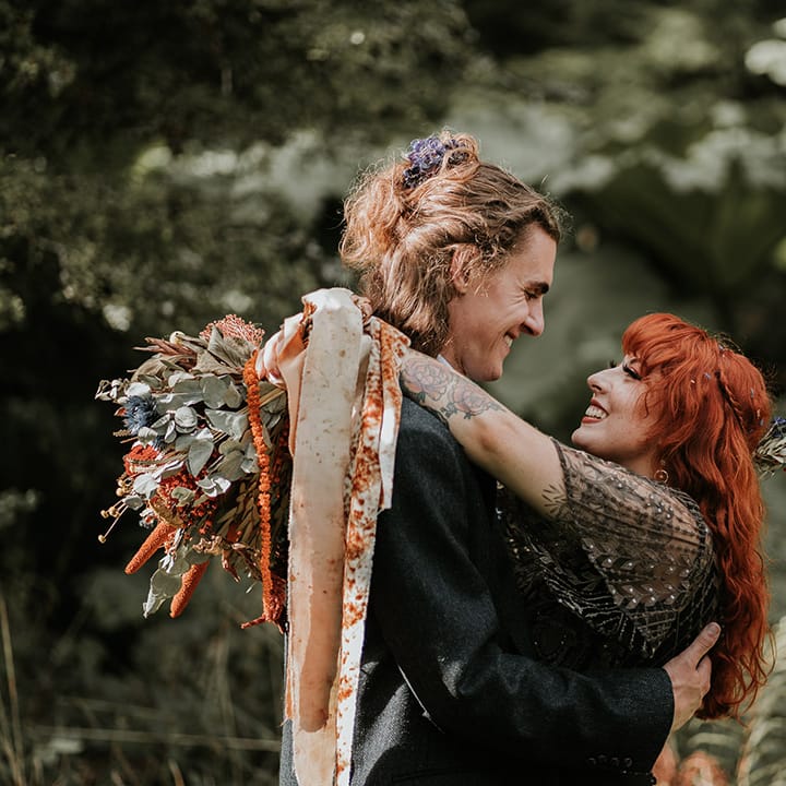 Bride in embellished short sleeve black wedding dress holding orange patterned ribbons embracing groom in dark grey suit 