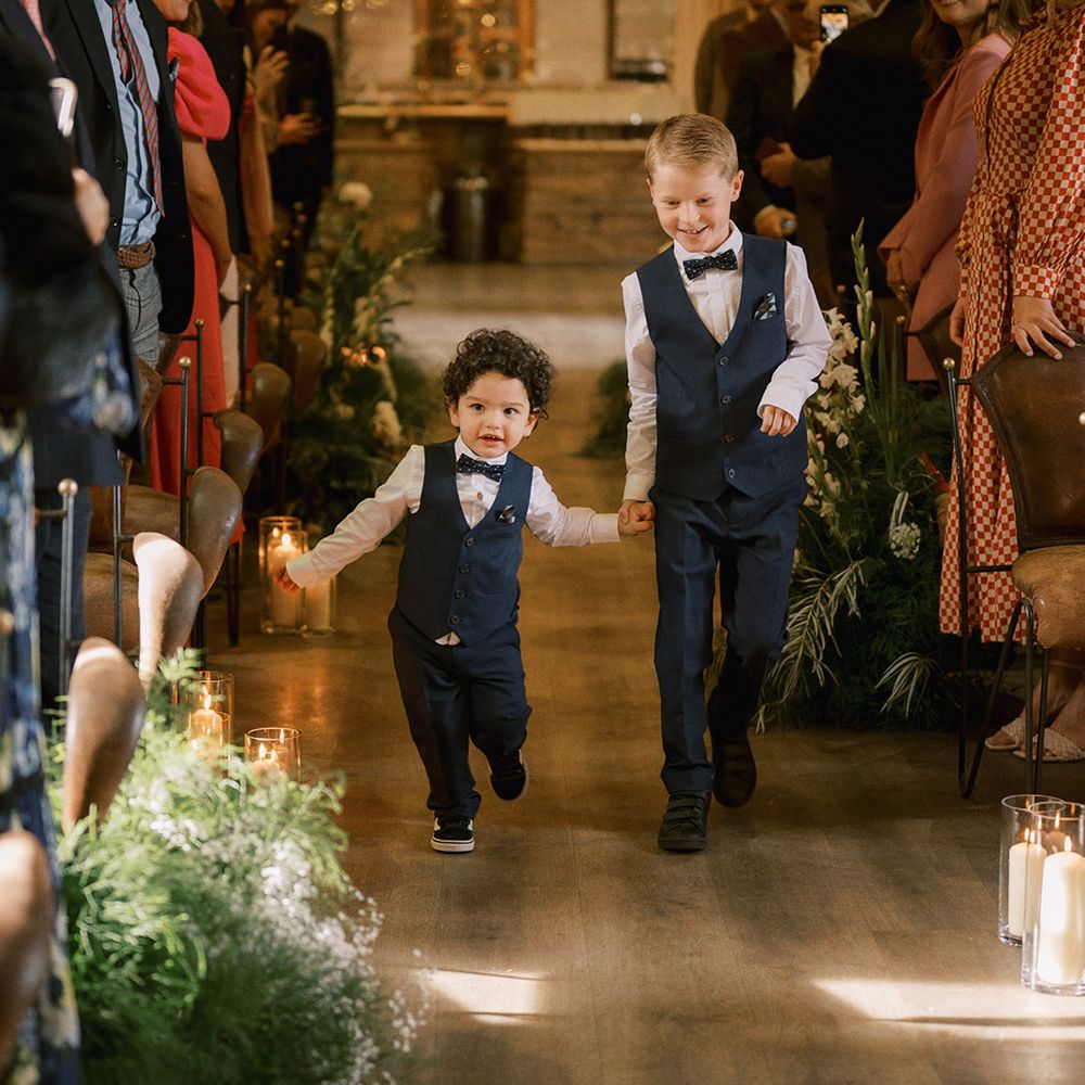 Page boys in navy waistcoat and bow tie walking down the aisle together with pillar candles in hurricane vases and white flowers decorating the aisles 