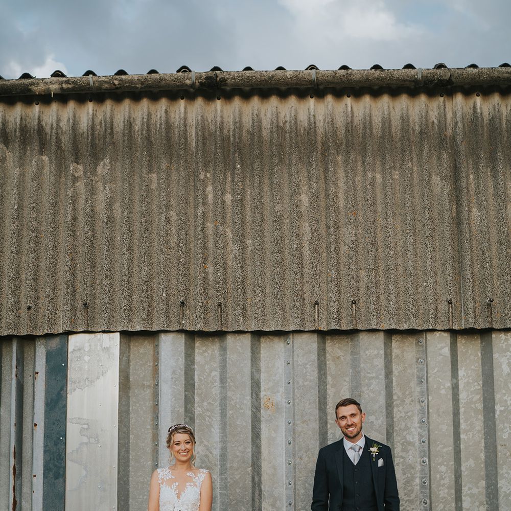 Bride in Dando London wedding dress and groom in dark blue suit stand in front of corrugated building 