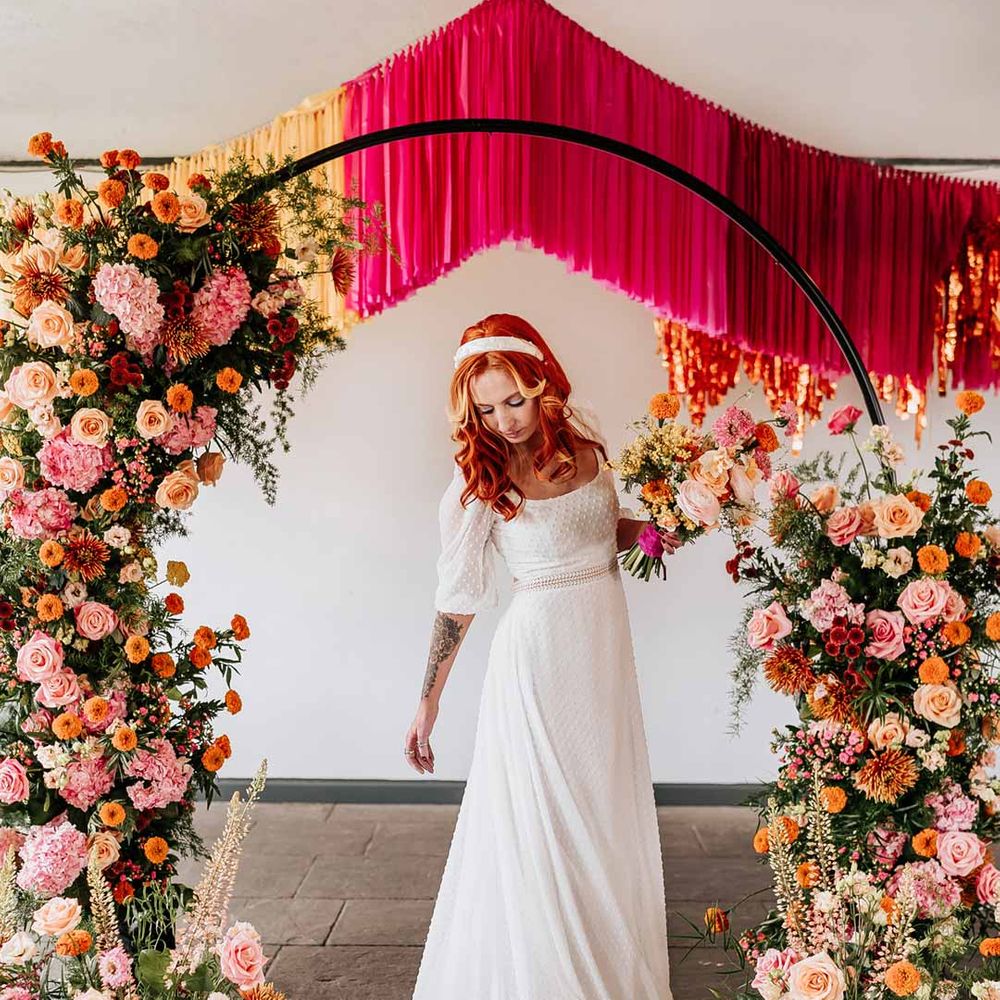 Bride in three quarter length puff sheer sleeve wedding dress with delicate polka dot overlay and square neck standing by circular floral arch with vibrant flowers and foliage attached and pink, red, yellow and metallic wedding streamers behind