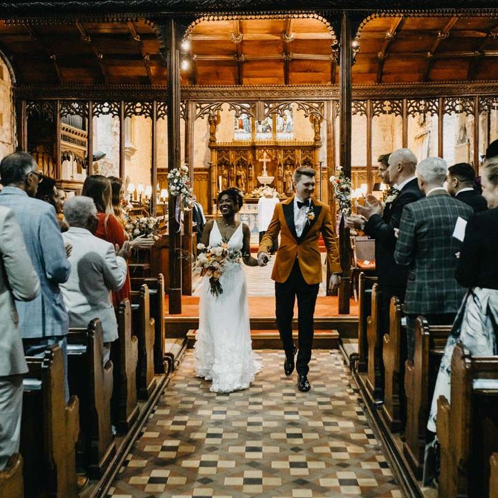 Groom in crushed burnt orange velvet grooms blazer, black bowtie and black suit trousers walking hand in hand with bride in v neck sleeveless wedding dress with 3D floral applique train holding blush and orange garden roses, poppies and carnations, dried flowers and eucalyptus wedding bouquet walking down the aisle at Brinsop Court wedding