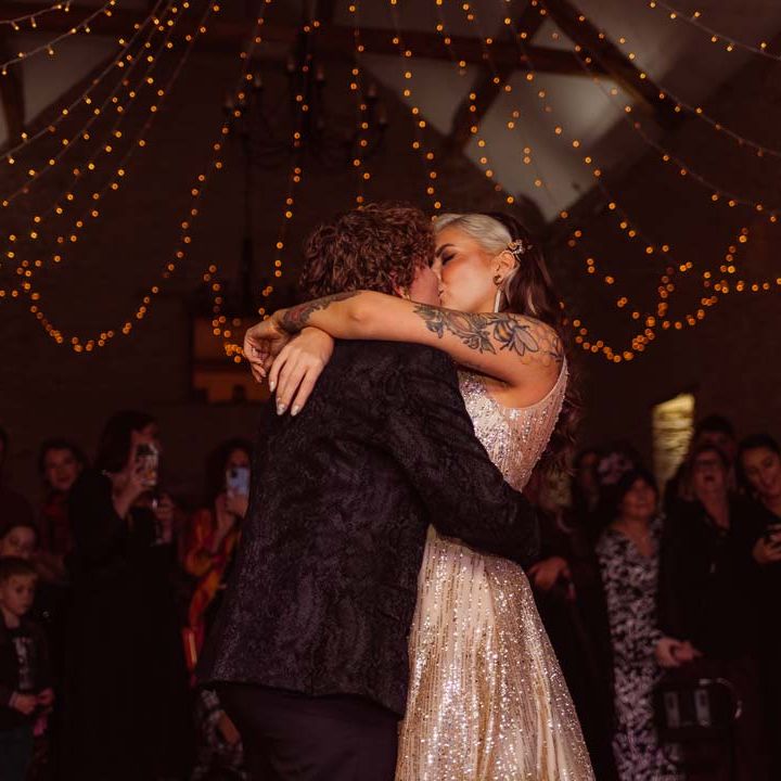 Bride in sleeveless deep v neck sparkly gold wedding dress and bride in dark velvet suit under fairy lights doing their wedding first dance at Kingscote Barn Tetbury wedding venue