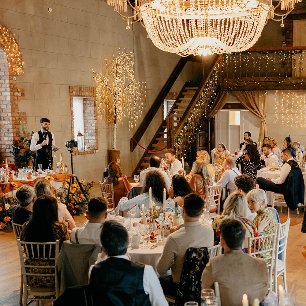 Wedding guests seated for the wedding breakfast with the groom reading out his wedding speech at NI wedding venue with chandelier 