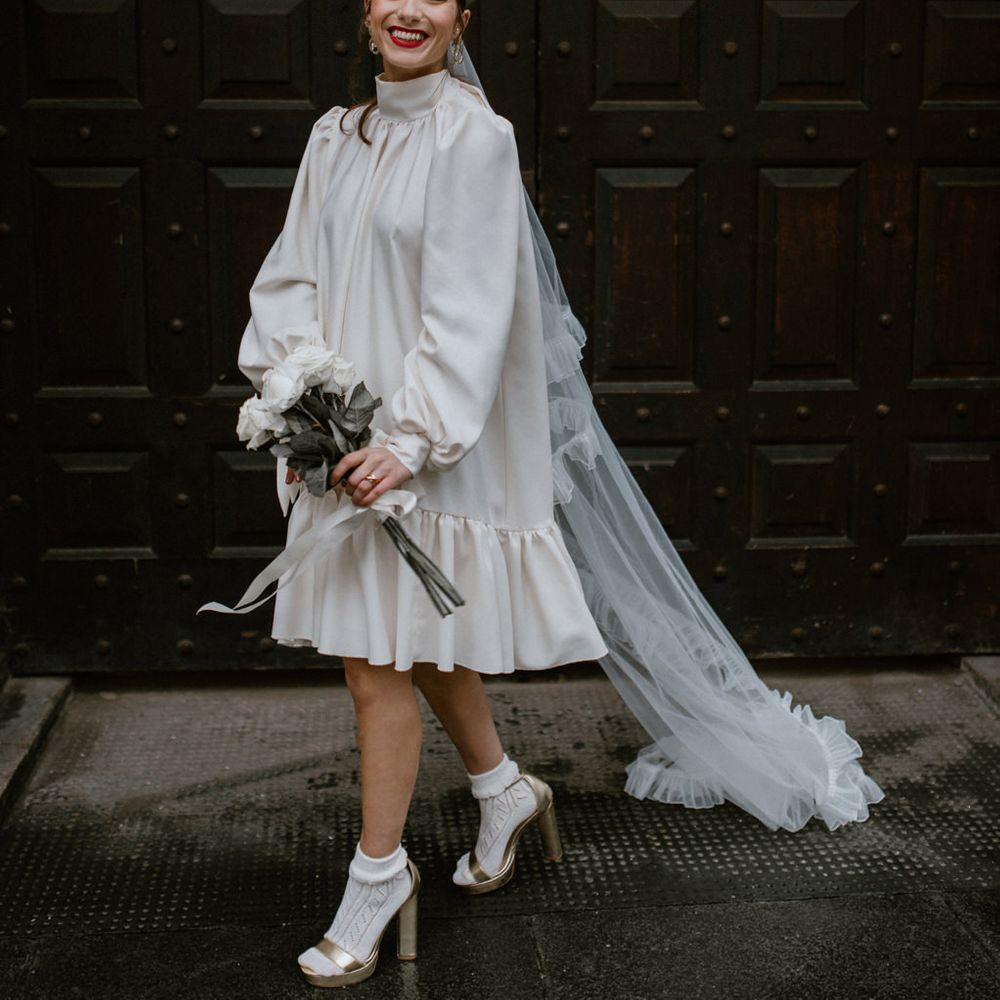 Stylish bride in a silk wedding dress with long sleeves and high neck wearing a long veil with ruffled edges, socks and gold platform Gucci sandals 