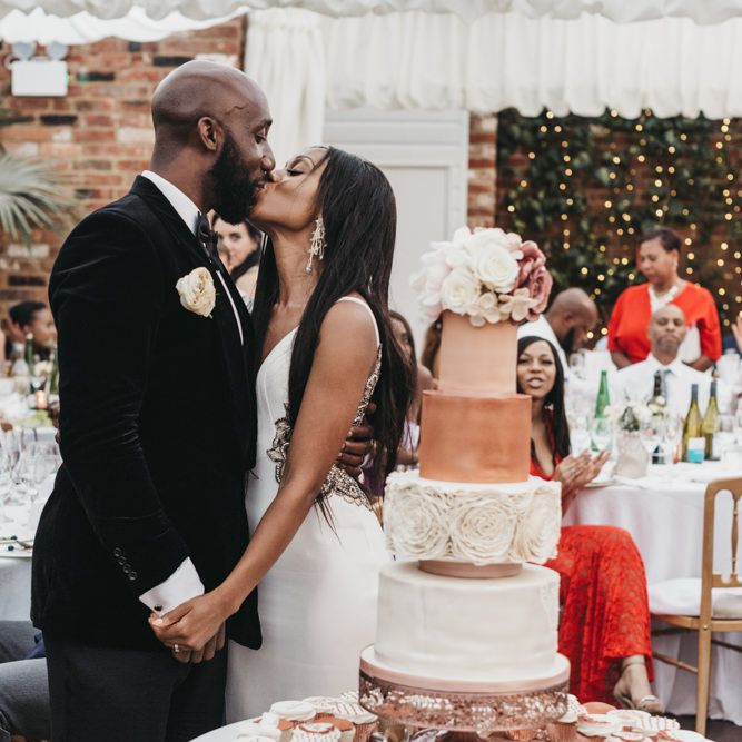 Black bride and groom kissing next to their white and rose gold wedding cake surrounded by individual cupcakes 