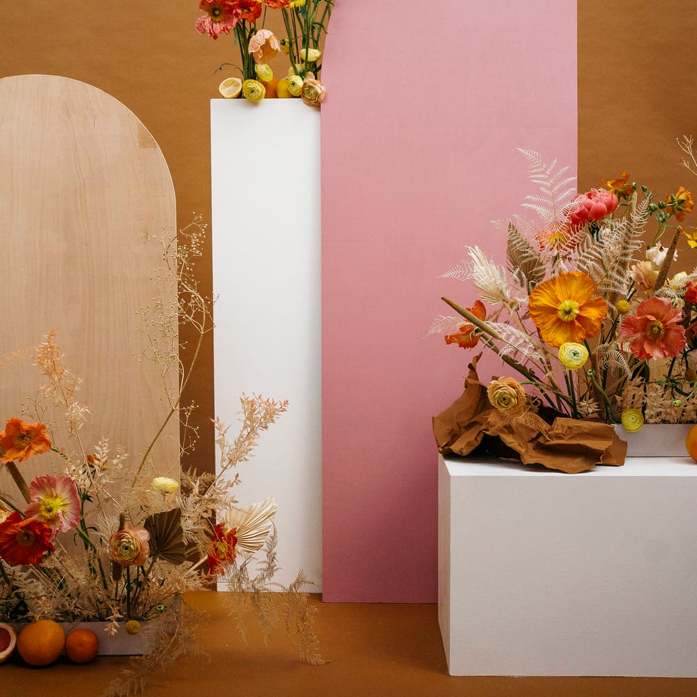 Wedding flowers on plinths including pink and yellow poppies, roses, dried palms, dried ferns and citrus fruit