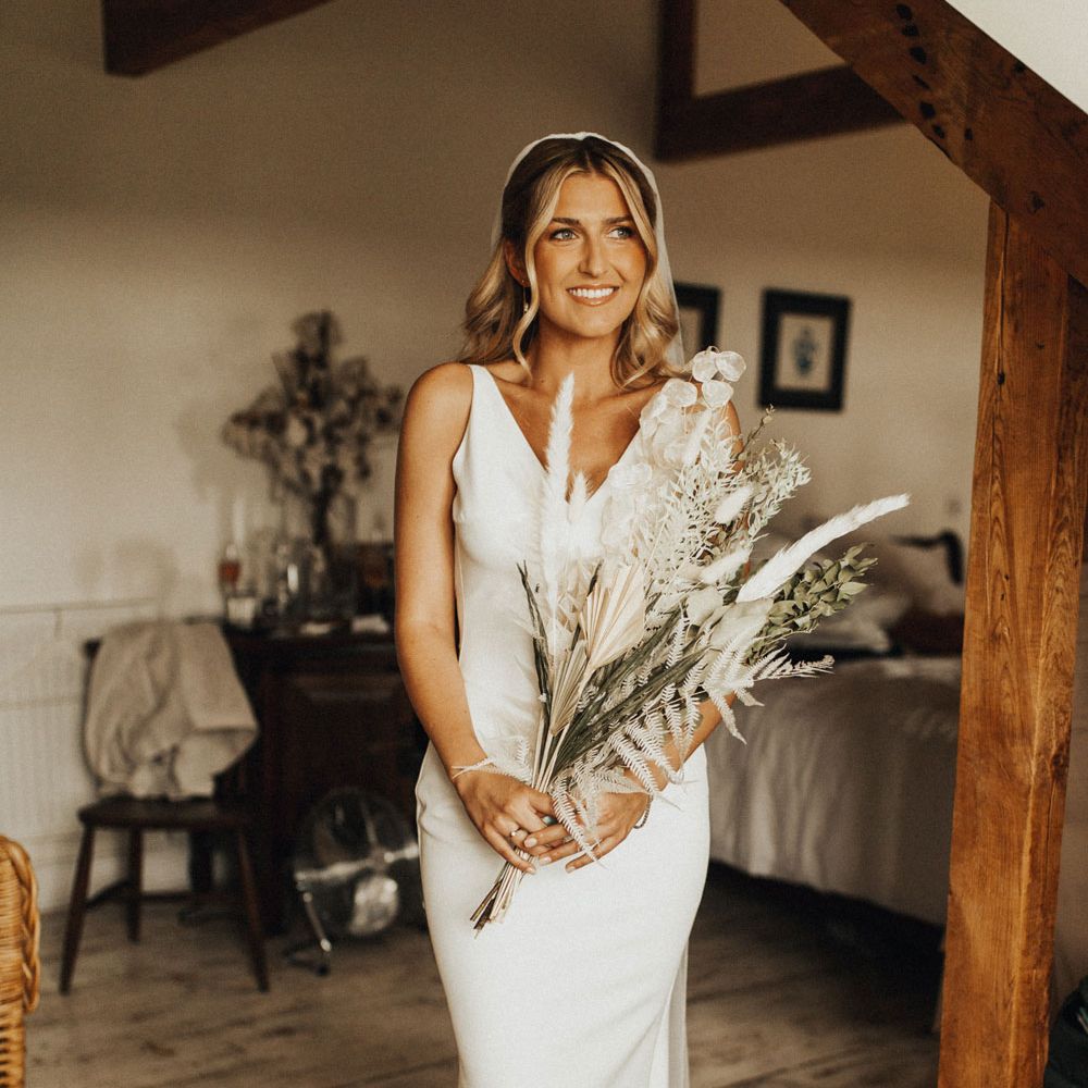 Smiling bride in V neck Made With Love wedding dress and veil holds white and green dried bridal bouquet in bedroom before wedding at Anran Devon