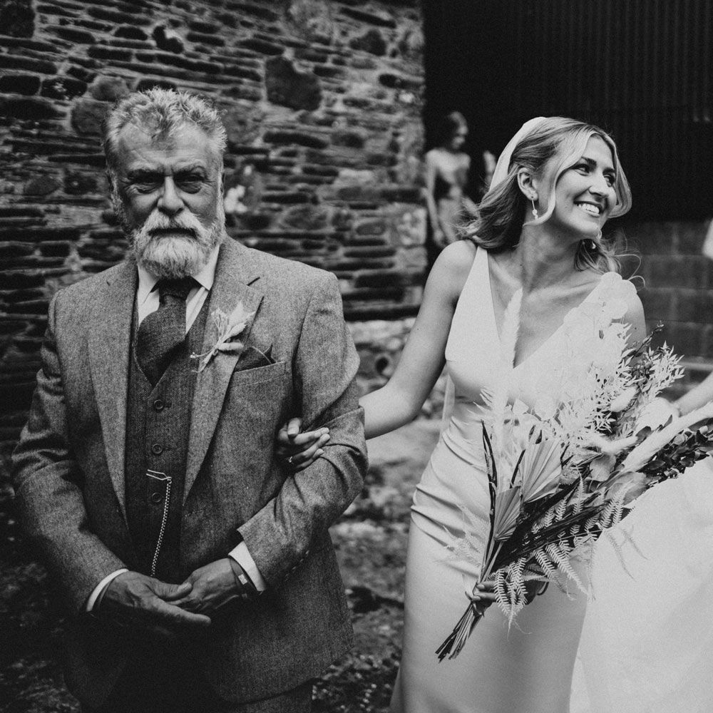 Smiling bride in V neck Made With Love wedding dress and veil holds dried bridal bouquet and arm of man in three piece suit as she walks to get married at Anran Devon