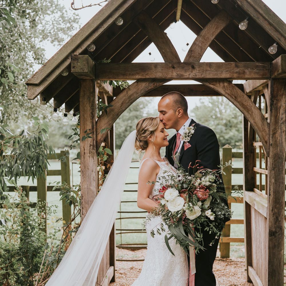 Bride in white lace Justin Alexander wedding dress with train and veil holding white, pink and green bridal bouquet stands by wooden arch as groom in dark three piece suit and pink tie kisses her on the forehead at Tythe Barn wedding with barn wedding flowers