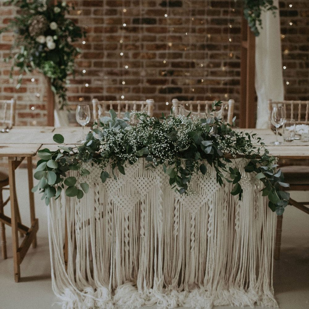 Wooden top table with macrame wedding decor, foliage drapery and bamboo chairs in front of wedding arch with cloth drapery and flower clouds for Isle of Wight wedding with macrame wedding decor
