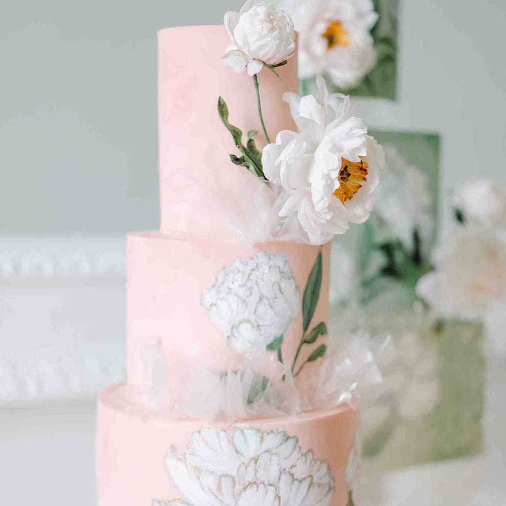 Light pink three tier wedding cake with white flower illustration on a glass cake stand 