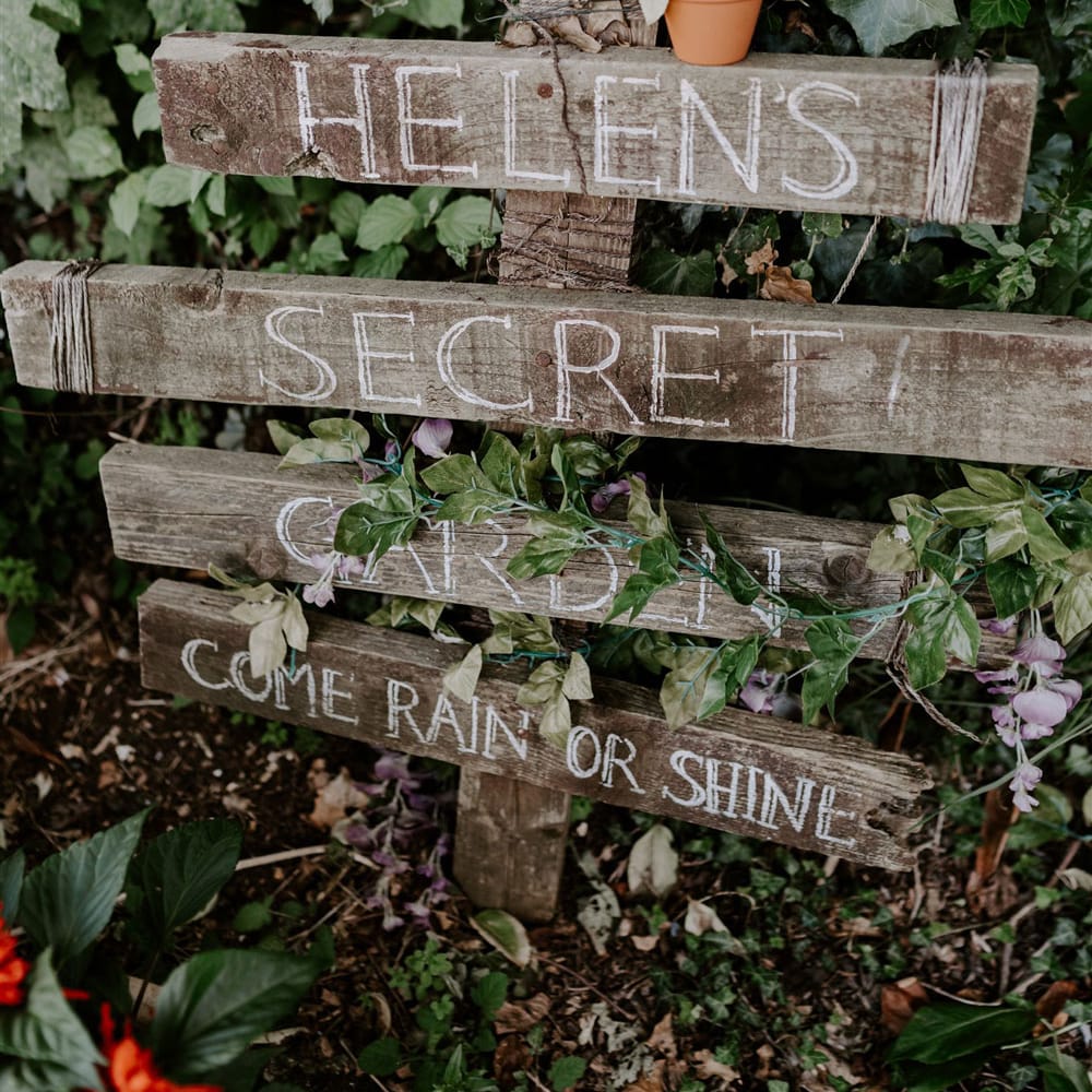 Wooden garden sign with four posts reading 'Helen's Secret Garden Come Rain or Shine' next to bush at DIY garden wedding