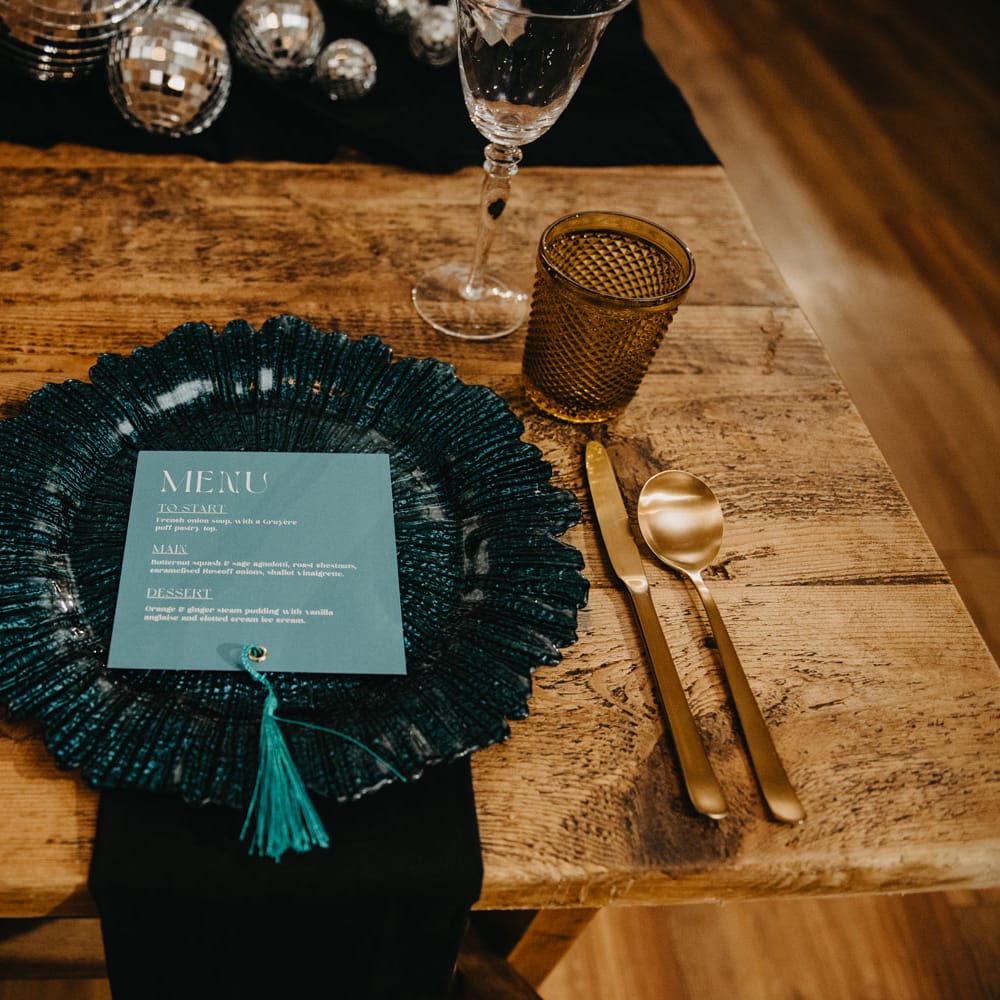 Blue glass plate with light blue wedding menu on wooden table with gold cutlery and orange glasses for rustic wedding breakfast