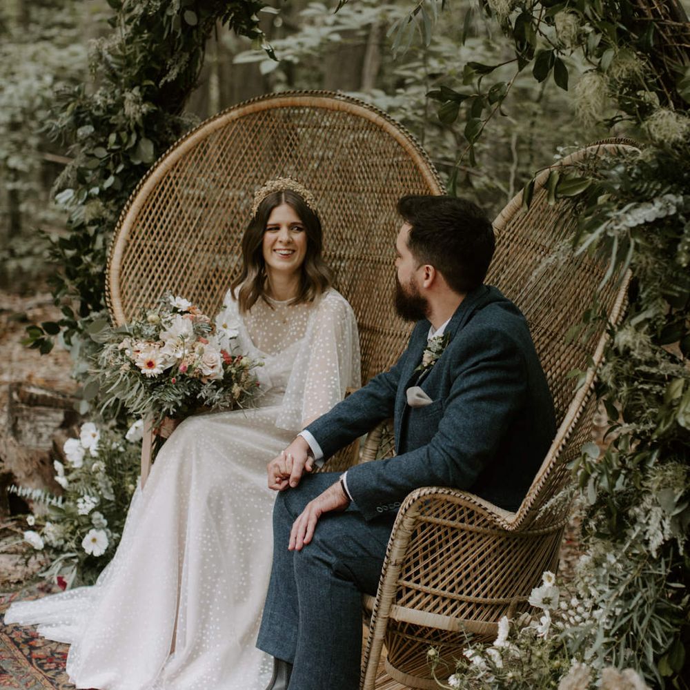 Bride & groom look lovingly at one another whilst they sit in wicker sweetheart chairs