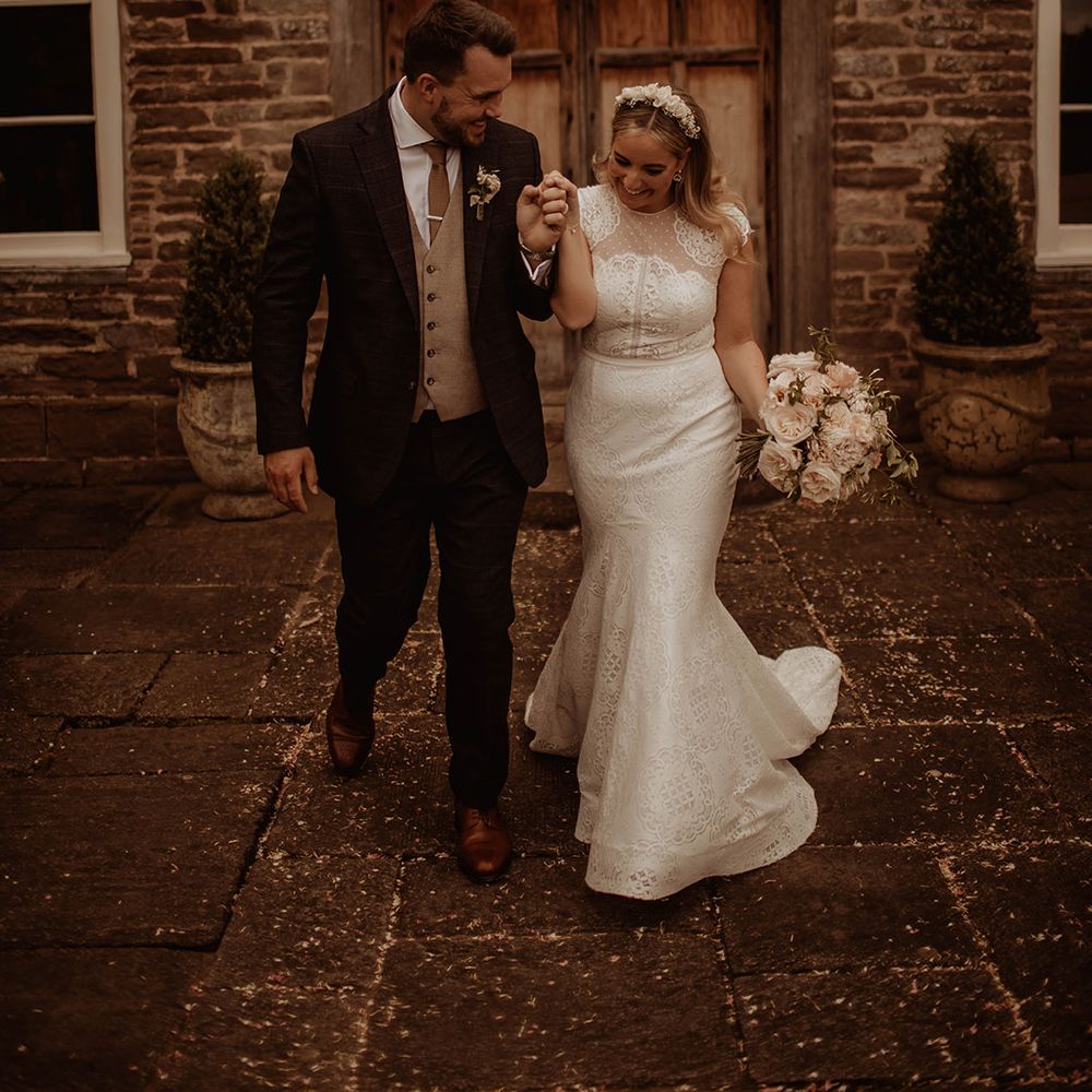 Bride in a lace cap sleeve wedding dress with mermaid skirt and groom in a three piece suit holding hands at their Dewsall Court wedding 