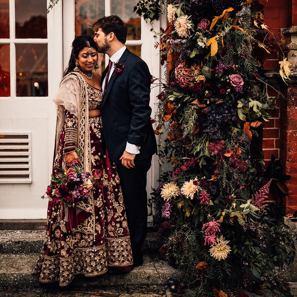 Bride in Indian Saree wedding dress stands with groom under their flower arch full of autumnal colours 