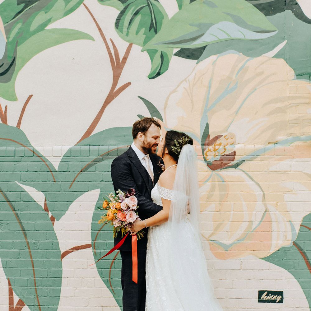 Bride in lace wedding dress with train and veil holding bouquet tied with red ribbon stands with groom in blue check suit in front of wall covered in street art in Liverpool