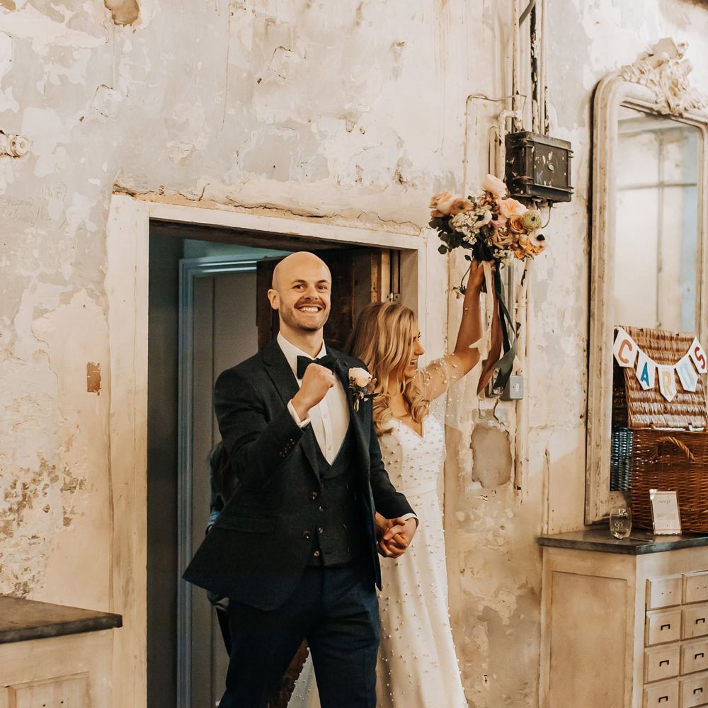 Groom in navy three piece tweed suit with bow tie walks into wedding reception with bride in white pearl wedding dress holding up pastel wedding bouquet