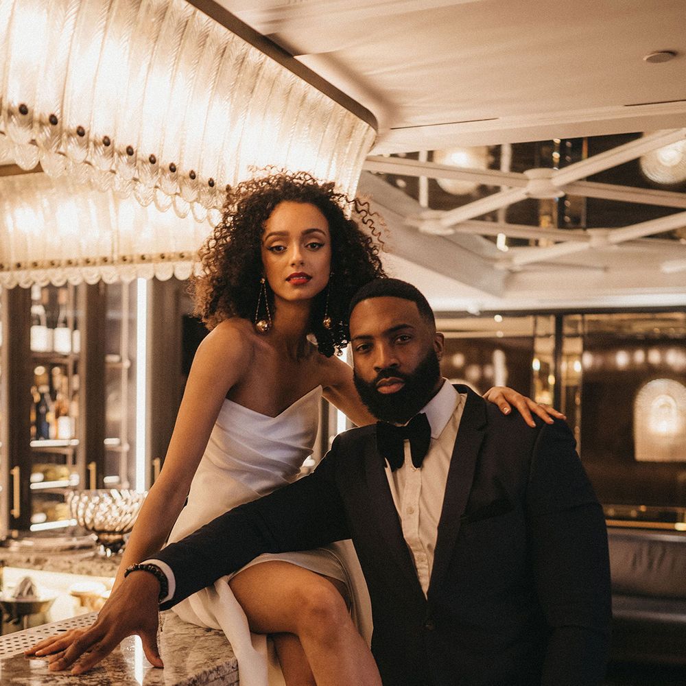 Black groom in a tuxedo standing at the bar with his Black bride in a strapless wedding dress with front slit sitting on the bar with naturally curly hair, red lipstick and winged eyeliner makeup 