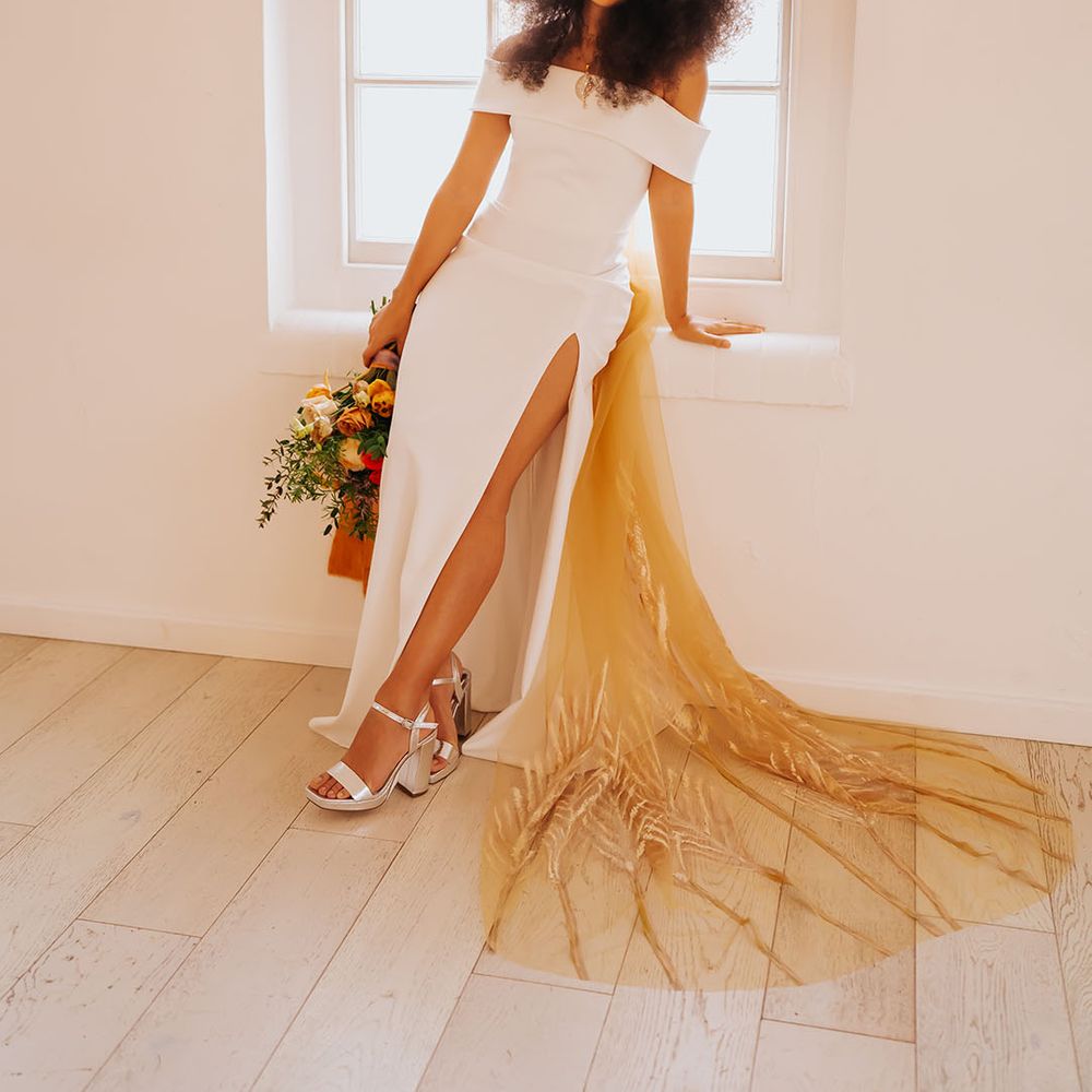 Black bride in an off the shoulder wedding dress with side slit wearing silver platform shoes and a patterned golden wedding veil
