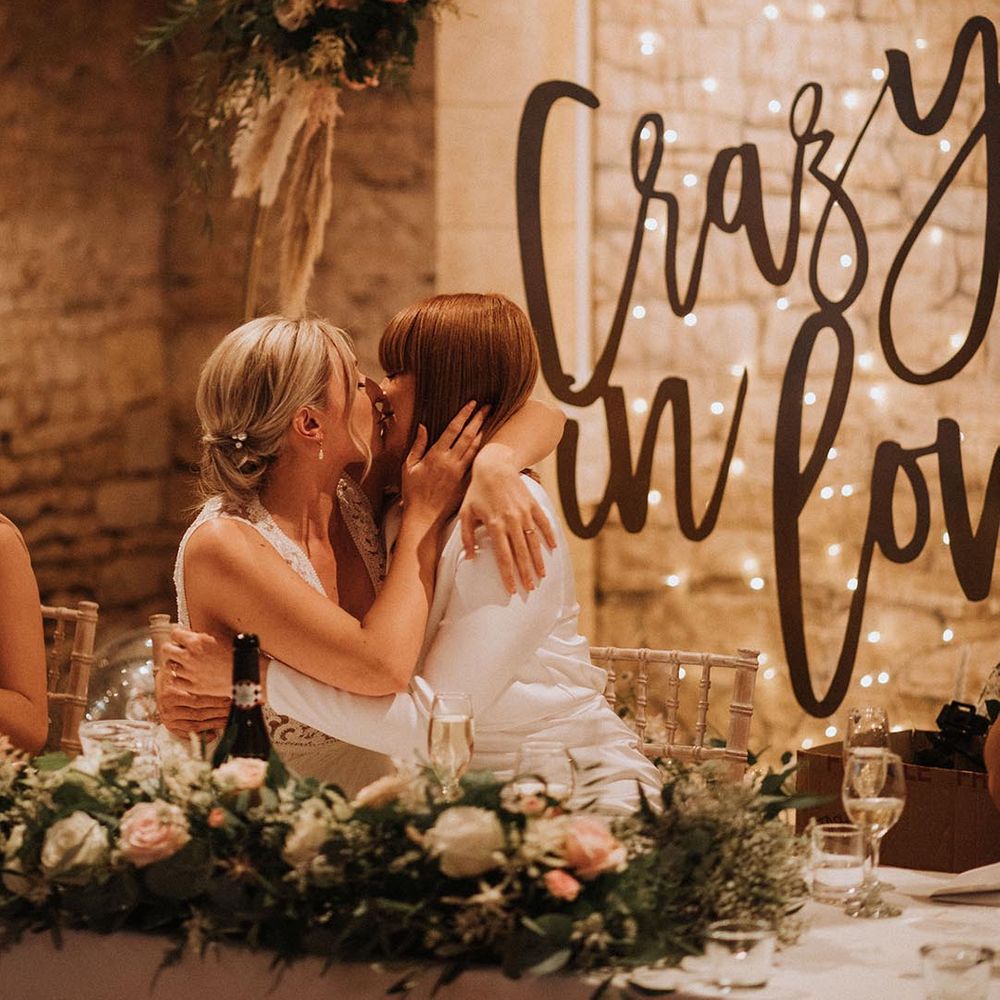 two brides kissing during the speeches at their rustic barn wedding with a giant Crazy In Love sign behind the top table 