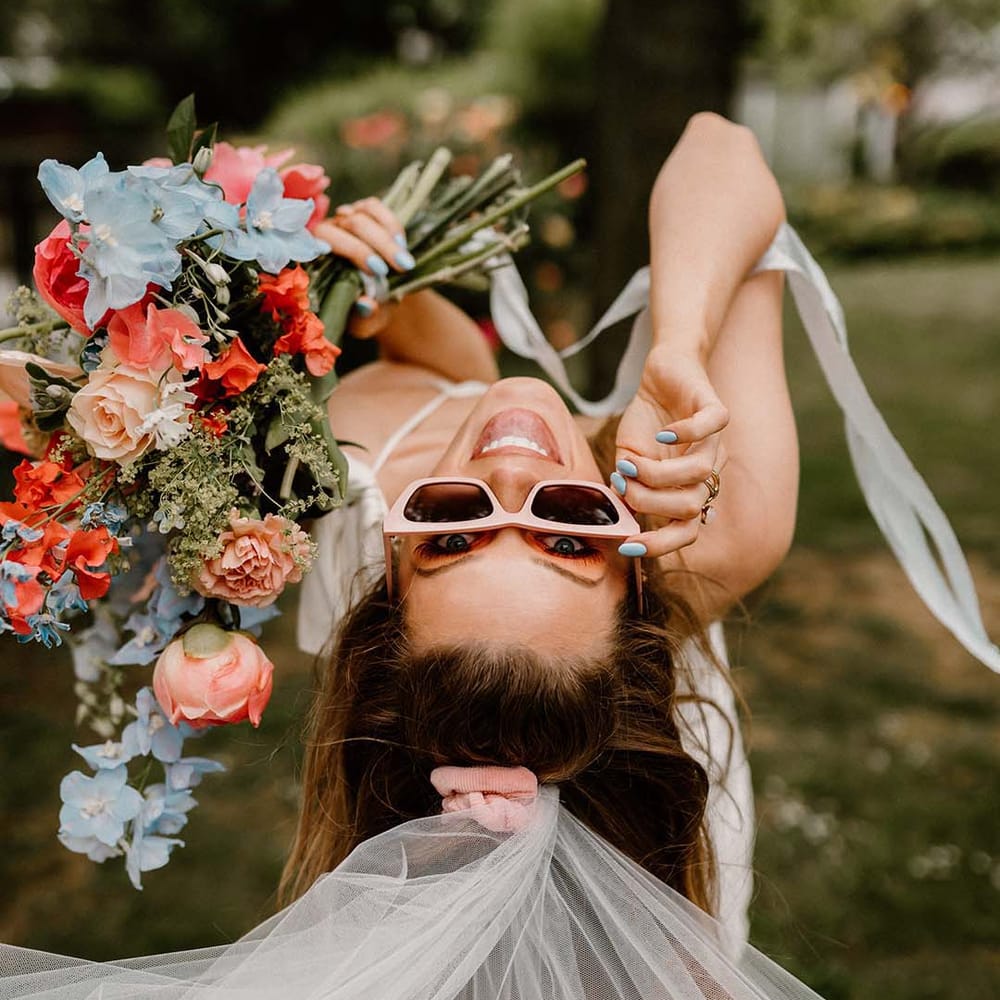 Bride in retro pink sunglasses holding a pink and blue wedding veil with peonies and anthuriums 